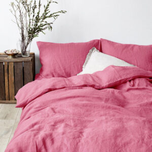Dans une chambre à coucher, un lit décoré d'une parure de lit en lin rose, avec coussins assortis et un coussin blanc, sur la gauche une plante verte se trouve dans un pot en bois