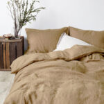 Dans une chambre à coucher, un lit décoré d'une parure de lit en lin beige foncé, avec coussins assortis et un coussin blanc, sur la gauche une plante verte se trouve dans un pot en bois