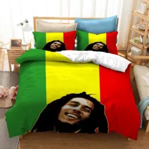 Dans une chambre le lit est décoré d'une parure à l'effigie de Bob Marley souriant, en fond les couleurs rouge, jaune et vert, avec la tête du chanteur bob marley en noir, les coussins sont assortis, à droite du lit il y a un banquette en bois avec les coussins blanc, et un meuble étagère en bois, à gauche du lit il y a une table de chevet en bois et des jouets