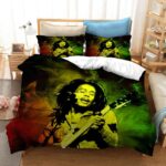 Dans une chambre le lit est décoré d'une parure à l'effigie de Bob Marley en train de jouer de la guitare, de couleur rouge, jaune et vert, avec la silhouette de chanteur bob marley en noir, les coussins sont assortis, à droite du lit il y a un banquette en bois avec les coussins blanc, et un meuble étagère en bois, à gauche du lit il y a une table de chevet en bois et des jouets