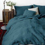 Dans une chambre à coucher, un lit décoré d'une parure de lit en lin verte, avec coussins assortis et un coussin rayé bleu et blanc, sur la gauche une plante verte se trouve une table de chevet en bois