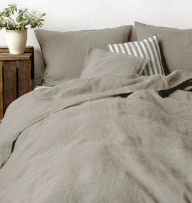 Dans une chambre à coucher, un lit décoré d'une parure de lit en lin khaki, avec coussins assortis et un coussin rayé gris et blanc, sur la gauche une plante verte se trouve une table de chevet en bois