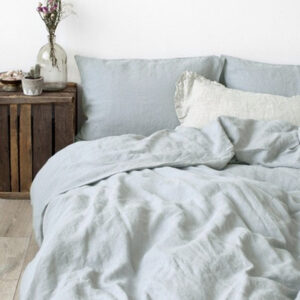 Dans une chambre à coucher, un lit décoré d'une parure de lit en lin bleu clair, avec coussins assortis et un coussin blanc, sur la gauche une plante verte se trouve une table de chevet en bois