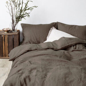Dans une chambre à coucher, un lit décoré d'une parure de lit en lin marron, avec coussins assortis et un coussin blanc, sur la gauche une plante verte se trouve une table de chevet en bois