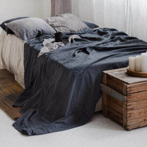 Dans une chambre le lit est décorée d'une belle parure de lit noire avec les coussins gris, au pied du lit se trouve une malle en bois avec des bougies dessus , le drap noir traine par terre