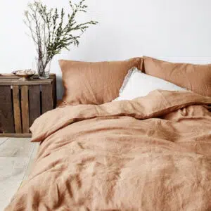 Dans une chambre à coucher, un lit décoré d'une parure de lit en lin couleur champagne, avec coussins assortis et un coussin blanc, sur la gauche une plante verte se trouve une table de chevet en bois