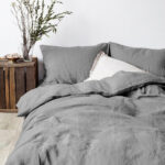Dans une chambre à coucher, un lit décoré d'une parure de lit en lin gris, avec coussins assortis et un coussin blanc, sur la gauche une plante verte se trouve une table de chevet en bois