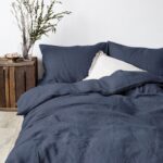 Dans une chambre à cuocher, un lit décoré d'une parure de lit en lin bleu foncé, avec ocussins assortis et un coussin blanc, sur la gauche une plante verte se trouve dans un pot en bois