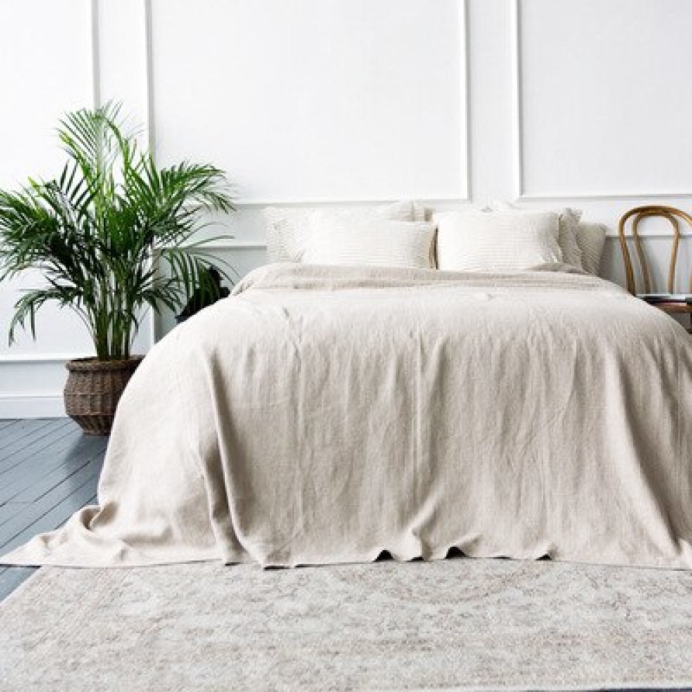 Dans une chambre un grand lit deux places est recouvert d'un belle parure de lin de couleur naturelle dont le drap plat plane sur le sol et le tapis de la chambre, à gauche un petit palmier, et à droite du lit un chaise en bois