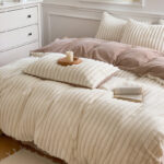 Parure de lit de couleur beige polaire avec livre ouvert dessus et un coussin avec un bougie sur un socle en bois