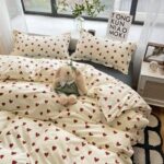 Parure de lit beige imprimés coeurs rouges sur un lit deux personnes. Une fenêtre en tête de lit et une table de chevet à droite.