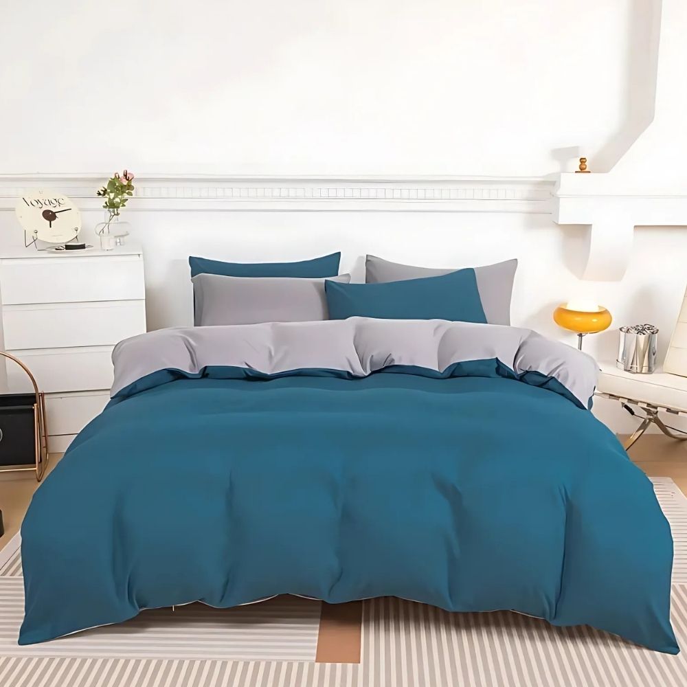 Parure de lit réversible bleue et grise sur un lit double avec le côté bleu sur l'extérieru. Un mur blanc en tête de lit, un meuble blanc sur la gauche. Des coussins assortis bleus et gris sur le lit.