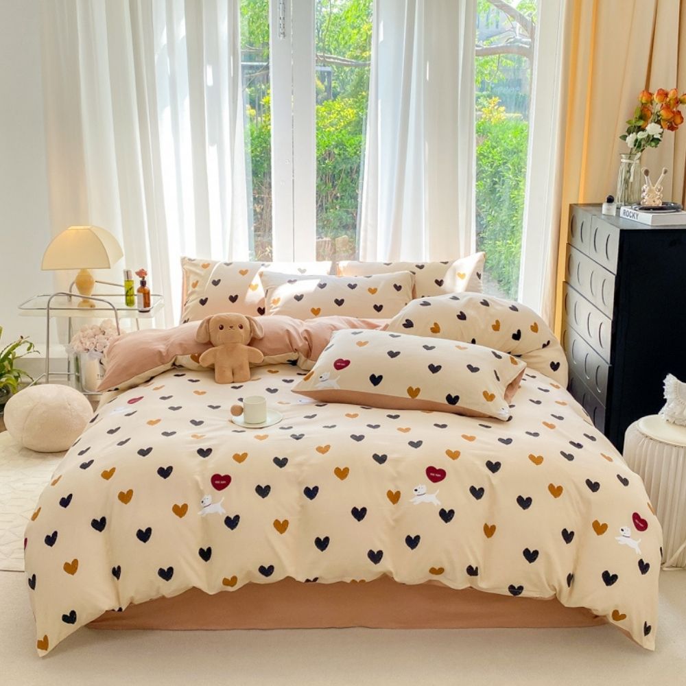 Parure de lit romantique beige avec des petits coeurs bleus et marrons et des petits chiens blancs. La parure est sur un lit deux places avec les taies d'oreillers assorties. Le lit se trouve devant une grande fenêtre avec des voilages blancs. A droite, un meuble noir, à gauche, une table de chevet en métal avec une lampe de chevet allumée.