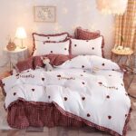 Parure de lit romantique bordeaux et blanche avec coeurs rouges sur un lit double. Plusieurs coussins assortis sont en haut du lit. De chaque côté du lit des tables de chevet rondes avec une lampe de chevet allumée à gauche, un cadre rectangulaire sur le mur. A droite, une décoration "love" sur la table de chevet et un rideau jaune à carreaux.