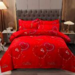 Parure de lit romantique rouge vif avec des coeurs et inscriptions brillantes. La housse de couette est assorties à deux coussins. L'ensemble se trouve sur un lit double dans une chambre avec une tête de lit matelassée marron. Une table de chevet de chaque côté du lit avec une lampe de chevet allumé.