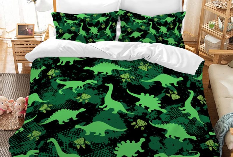 Dans une chambre le lit est décoré d'une parure avec plusieurs motifs de dinosaures verts, le reste de la couverture est noire et le verso est blanc, les coussins sont assortis, à droite du lit il y a un banquette en bois avec les coussins blanc, et un meuble étagère en bois, à gauche du lit il y a une table de chevet en bois et des jouets