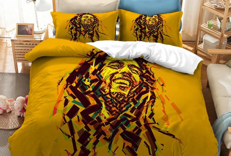 Dans une chambre le lit est décoré d'une parure à l'effigie de Bob Marley dessiné, de couleur orange, avec le visage du chanteur bob marley dessiné avec des petites touches oranges, les coussins sont assortis, à droite du lit il y a un banquette en bois avec les coussins blanc, et un meuble étagère en bois, à gauche du lit il y a une table de chevet en bois et des jouets