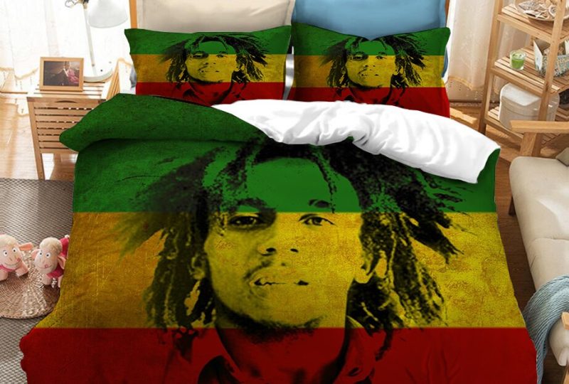 Dans une chambre le lit est décoré d'une parure à l'effigie de Bob Marley jeune avec les dreadlocks court, l'ensemble est de couleur rouge, jaune et vert, avec le visage de bob marley en noir, les coussins sont assortis, à droite du lit il y a un banquette en bois avec les coussins blanc, et un meuble étagère en bois, à gauche du lit il y a une table de chevet en bois et des jouets