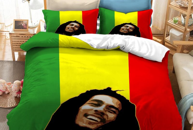 Dans une chambre le lit est décoré d'une parure à l'effigie de Bob Marley souriant, en fond les couleurs rouge, jaune et vert, avec la tête du chanteur bob marley en noir, les coussins sont assortis, à droite du lit il y a un banquette en bois avec les coussins blanc, et un meuble étagère en bois, à gauche du lit il y a une table de chevet en bois et des jouets