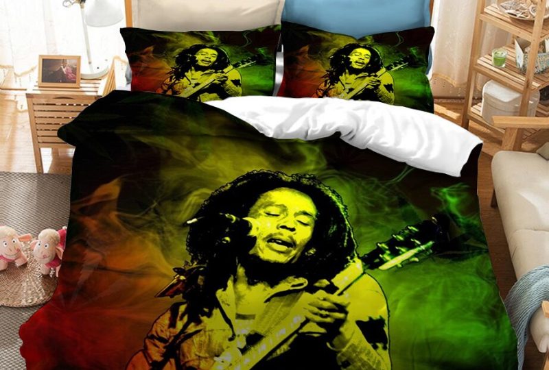 Dans une chambre le lit est décoré d'une parure à l'effigie de Bob Marley en train de jouer de la guitare, de couleur rouge, jaune et vert, avec la silhouette de chanteur bob marley en noir, les coussins sont assortis, à droite du lit il y a un banquette en bois avec les coussins blanc, et un meuble étagère en bois, à gauche du lit il y a une table de chevet en bois et des jouets