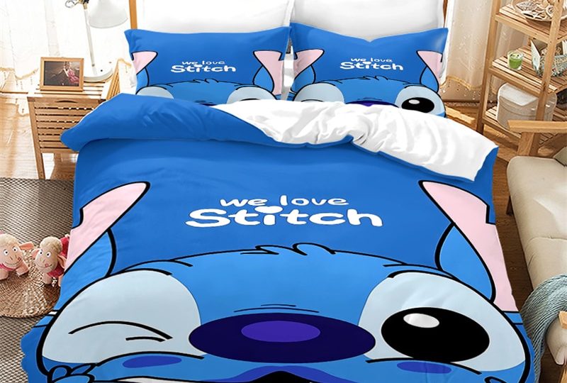 Dans une chambre le lit est décoré d'une parure à l'effigie du petit personnage Stitch bleu, les coussins sont assortis, à droite du lit il y a un banquette en bois avec les coussins blanc, et un meuble étagère en bois, à gauche du lit il y a une table de chevet en bois et des jouets