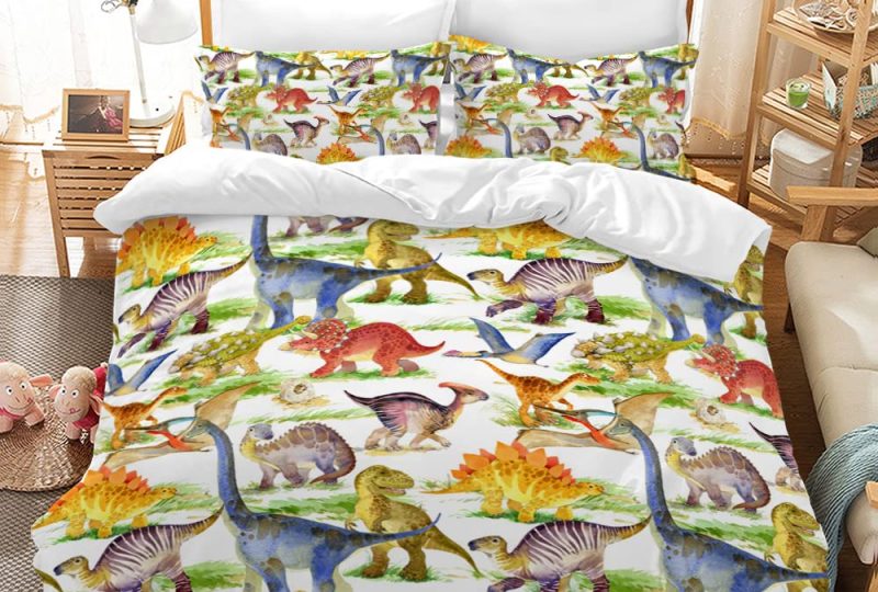Dans une chambre le lit est décoré d'une parure avec plusieurs de dinosaures en couleurs, le reste de la couverture est blanche, les coussins sont assortis, à droite du lit il y a un banquette en bois avec les coussins blanc, et un meuble étagère en bois, à gauche du lit il y a une table de chevet en bois et des jouets