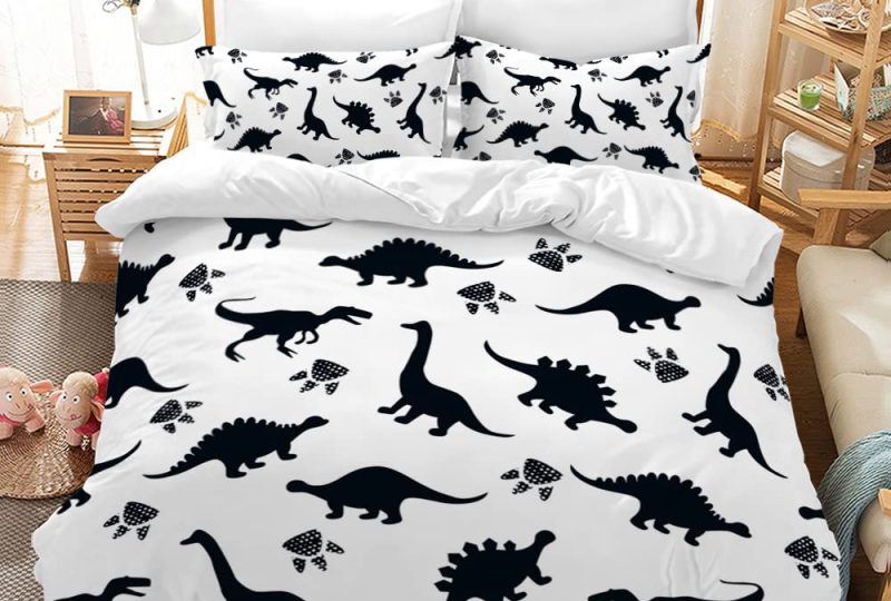 Dans une chambre le lit est décoré d'une parure avec plusieurs motifs de dinosaures noirs, le reste de la couverture est blanche, les coussins sont assortis, à droite du lit il y a un banquette en bois avec les coussins blanc, et un meuble étagère en bois, à gauche du lit il y a une table de chevet en bois et des jouets