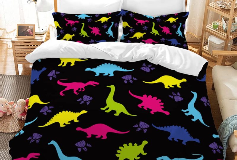 Dans une chambre le lit est décoré d'une parure avec plusieurs motifs de dinosaures multicolores, le reste de la couverture est noire et le verso est blanc, les coussins sont assortis, à droite du lit il y a un banquette en bois avec les coussins blanc, et un meuble étagère en bois, à gauche du lit il y a une table de chevet en bois et des jouets
