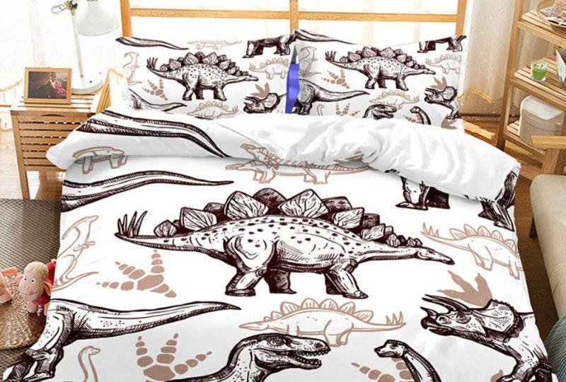 Dans une chambre le lit est décoré d'une parure avec plusieurs de dinosaures dessinés, le reste de la couverture est blanche, les coussins sont assortis, à droite du lit il y a un banquette en bois avec les coussins blanc, et un meuble étagère en bois, à gauche du lit il y a une table de chevet en bois et des jouets