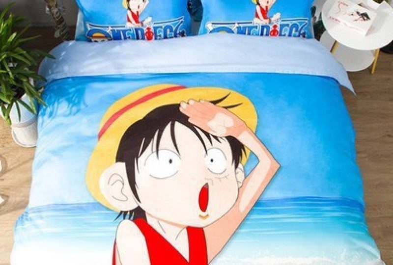 Parure de lit One piece multicolore où l'on voit le personnage Monkey D. Luffy sur une île déserte en plein soleil, cette parure est installée sur un lit dans une chambre avec une table de chevet ronde à droite et une plante verte à gauche