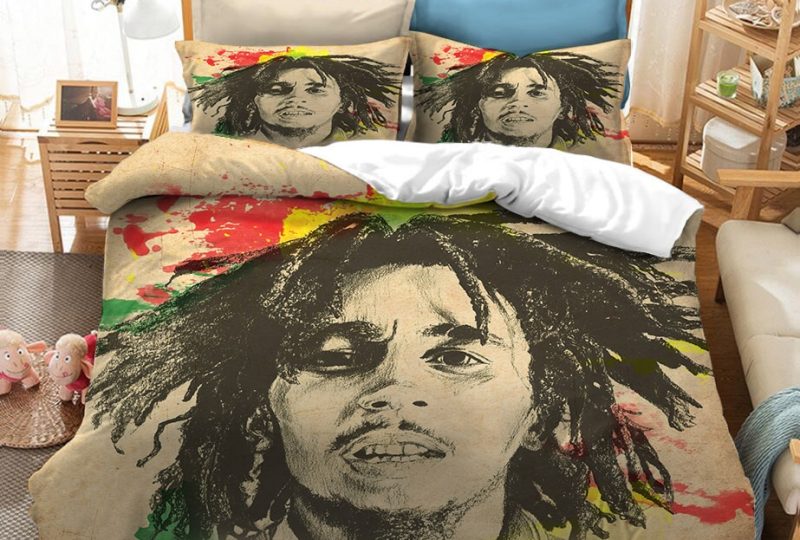 Dans une chambre le lit est décoré d'une parure à l'effigie de Bob Marley dessiné comme au fusin, de couleur beige avec des touches de couleurs, avec le visage du chanteur bob marley dessiné au fusin, les coussins sont assortis, à droite du lit il y a un banquette en bois avec les coussins blanc, et un meuble étagère en bois, à gauche du lit il y a une table de chevet en bois et des jouets