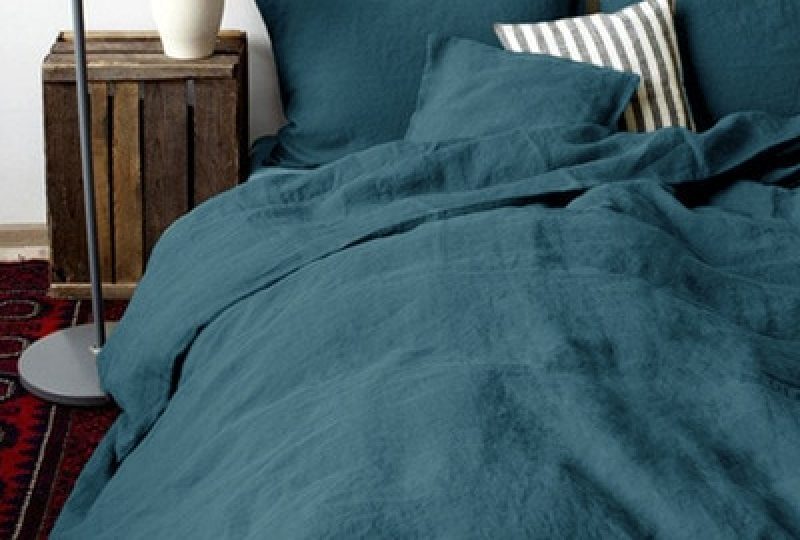 Dans une chambre à coucher, un lit décoré d'une parure de lit en lin verte, avec coussins assortis et un coussin rayé bleu et blanc, sur la gauche une plante verte se trouve une table de chevet en bois