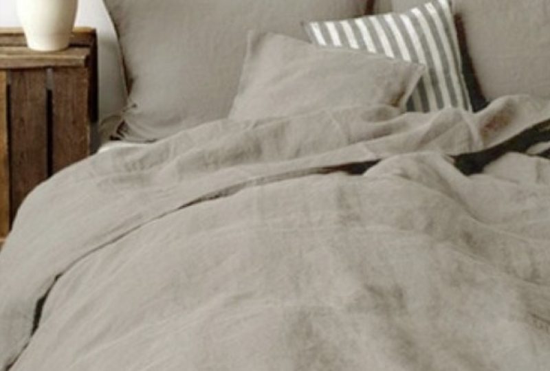 Dans une chambre à coucher, un lit décoré d'une parure de lit en lin khaki, avec coussins assortis et un coussin rayé gris et blanc, sur la gauche une plante verte se trouve une table de chevet en bois