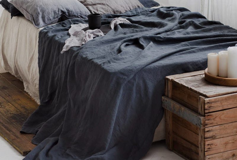 Dans une chambre le lit est décorée d'une belle parure de lit noire avec les coussins gris, au pied du lit se trouve une malle en bois avec des bougies dessus , le drap noir traine par terre
