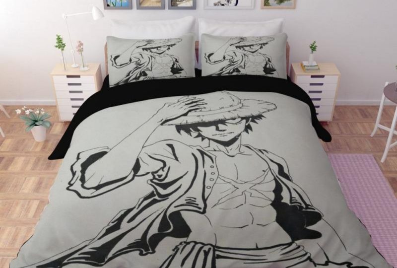 Dans une chambre, installée sur le lit et les oreillers, une parure complète à l'effigie du manga One Piece avec l'image du personnage Monkey D. Luffy en noir et blanc sur fond gris , de chaque côté du lit il y a une table de chevet blanche et carrée, au dessus du lit des affiches sont accrochées au mur et un tapis violet se trouve au sol
