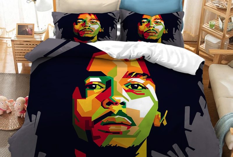 Dans une chambre le lit est décoré d'une parure à l'effigie de Bob Marley dessiné en style moderne, le fond est de couleur grise, avec le visage du chanteur bob marley dessiné avec des petites touches de couleurs, les coussins sont assortis, à droite du lit il y a un banquette en bois avec les coussins blanc, et un meuble étagère en bois, à gauche du lit il y a une table de chevet en bois et des jouets