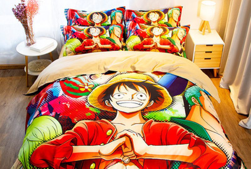 Parure de lit One piece multicolore où l'on voit le personnage Monkey D. Luffy les mains jointes, cette parure est installée sur un lit dans une chambre avec une table de chevet ronde à gauche et une table de chevet carrée avec une lampe dessus à droite
