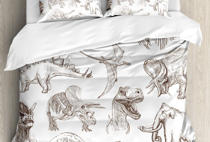 Dans une chambre le lit est décoré d'une parure avec plusieurs de dinosaures dessinés finement, le reste de la couverture est blanche, les coussins sont assortis, à droite du lit il y a un banquette en bois avec les coussins blanc, et un meuble étagère en bois, à gauche du lit il y a une table de chevet en bois et des jouets