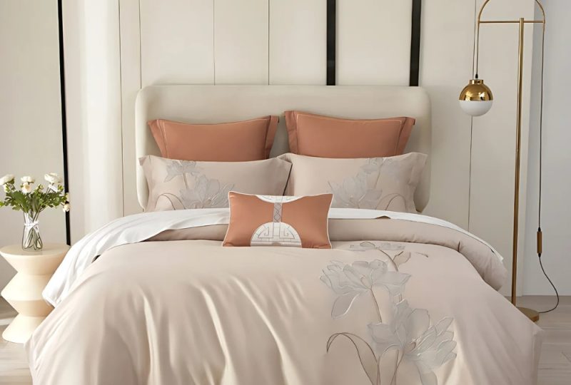 Parure de lit style asiatique beige sur un lit deux places avec le coussins assortis. Le mur côté tête de lit est blanc, une lampe portée à droite et un petit élément à gauche.