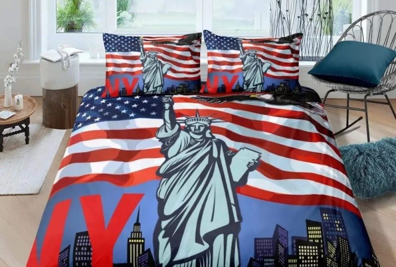 Parure de lit New York avec la Statue de la Liberté devant et des drapeaux américains derrière, les lettres N et Y en rouge sur la gauche. Deux coussins assortis sur un lit double. Au fond, une grande fenêtre et une chaise en métal sur la droite.