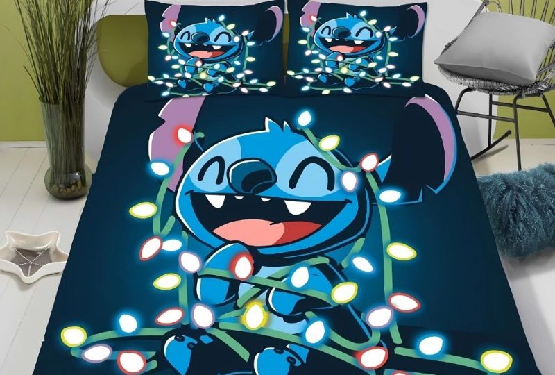 Parure de lit Stitch originale bleu marine avec Stitch souriant enroulé dans une guirlande lumineuse. Deux coussins sont assortis. Sur un lit double avec une tête de lit blanche, une chaise en métal sur la droite et de la verdure à gauche dans un vase.
