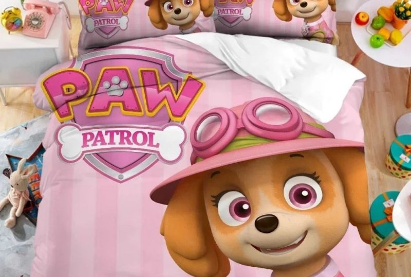 Parure de lit Pat'Patrouille rose avec Stella sur le coin droit et le logo de la Pat'Patrouille en haut à gauche. Les deux coussins sont assortis à la housse de couette. Sur un lit double dans une chambre avec une table de chevet à gauche et une petite table ronde blanche à droite.