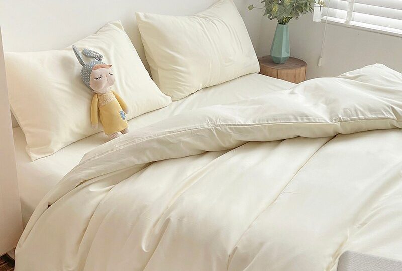 Parure de lit beige avec une poupée jaune posée sur le coussin avec une table basse en bois avec un vase de couleur bleu posé dessus