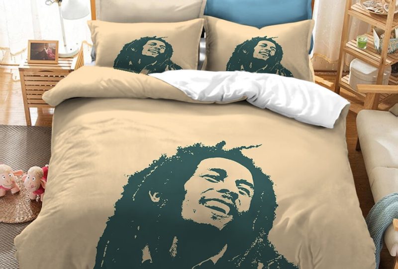 Dans une chambre le lit est décoré d'une parure à l'effigie de Bob Marley dessiné, de couleur beige, avec le visage du chanteur bob marley, les coussins sont assortis, à droite du lit il y a un banquette en bois avec les coussins blanc, et un meuble étagère en bois, à gauche du lit il y a une table de chevet en bois et des jouets