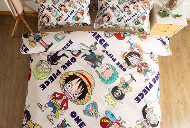 Parure de lit One piece multicolore avec plusieurs motifs des personnage de One Piece et des écritures, avec Monkey D. Luffy en élément central, sur la droite du lit il y a une table de chevet
